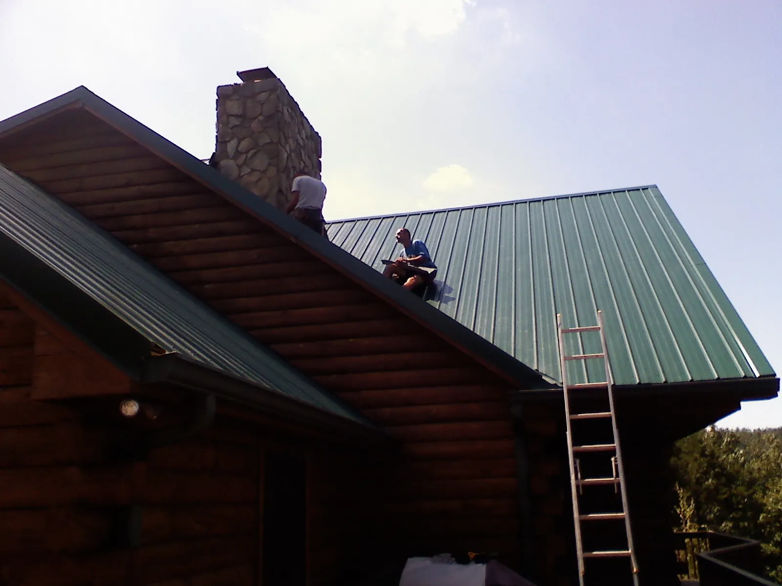 Roofers installing green metal panels beside a stone chimney on a log home in Parma